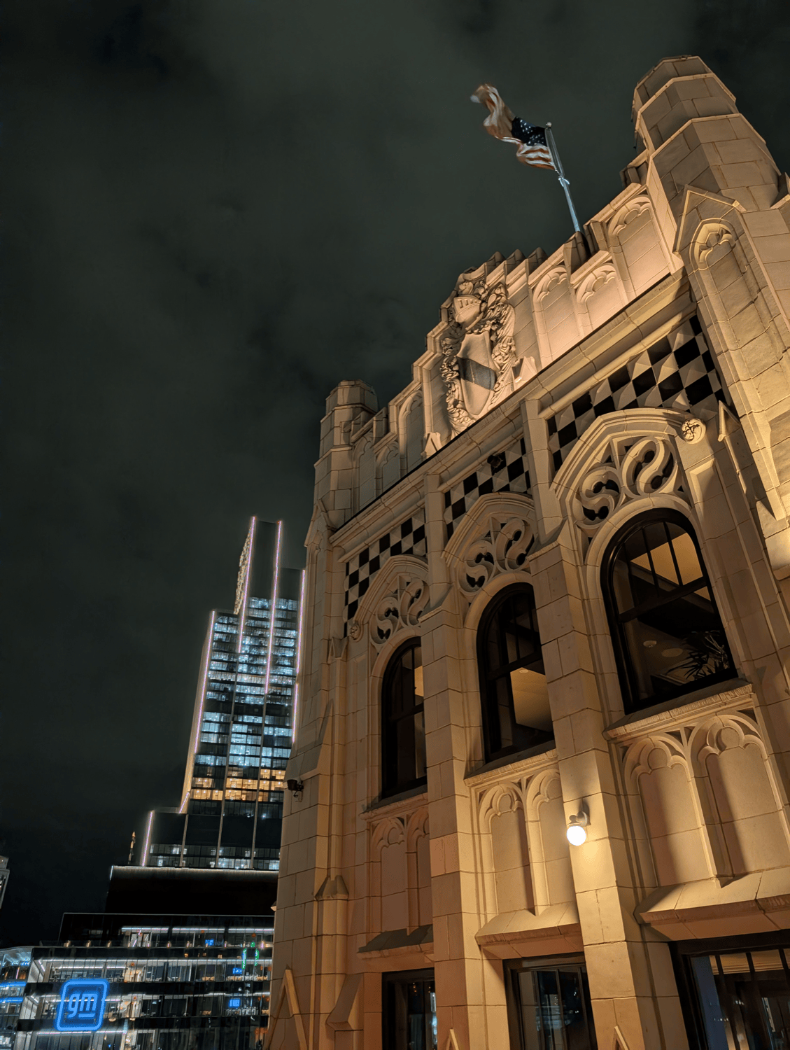 A Gothic building lit at night with a United States flag and the GM Renaissance Center glowing behind