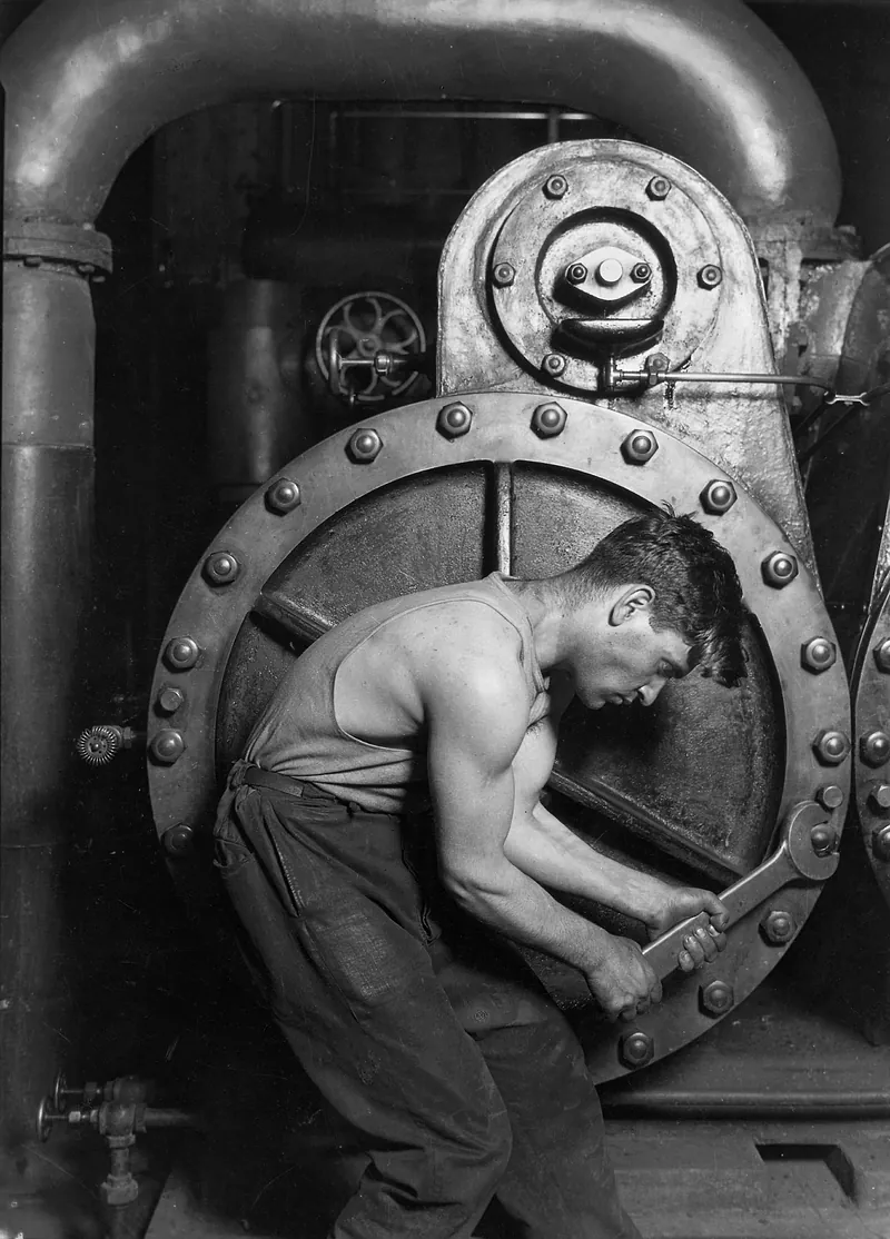 A photograph of a young mechanic bent into the circular housing of a large steam pump, both hands on a wrench, his back and arms taut with effort.